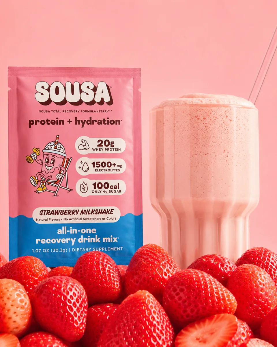 SOUSA Strawberry Milkshake protein and hydration recovery drink sachet with pink branding, positioned in front of a milkshake and strawberries